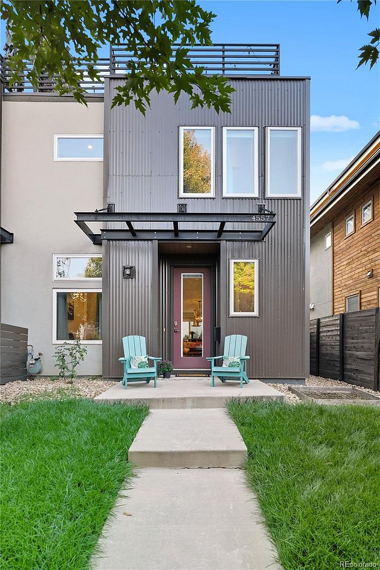 The image showcases the front exterior of a modern, two-story home. The facade combines corrugated metal siding with a light gray stucco, creating a contemporary aesthetic. A small, well-manicured lawn and a concrete walkway lead to the front door, flanked by two light blue Adirondack chairs, adding a welcoming touch.