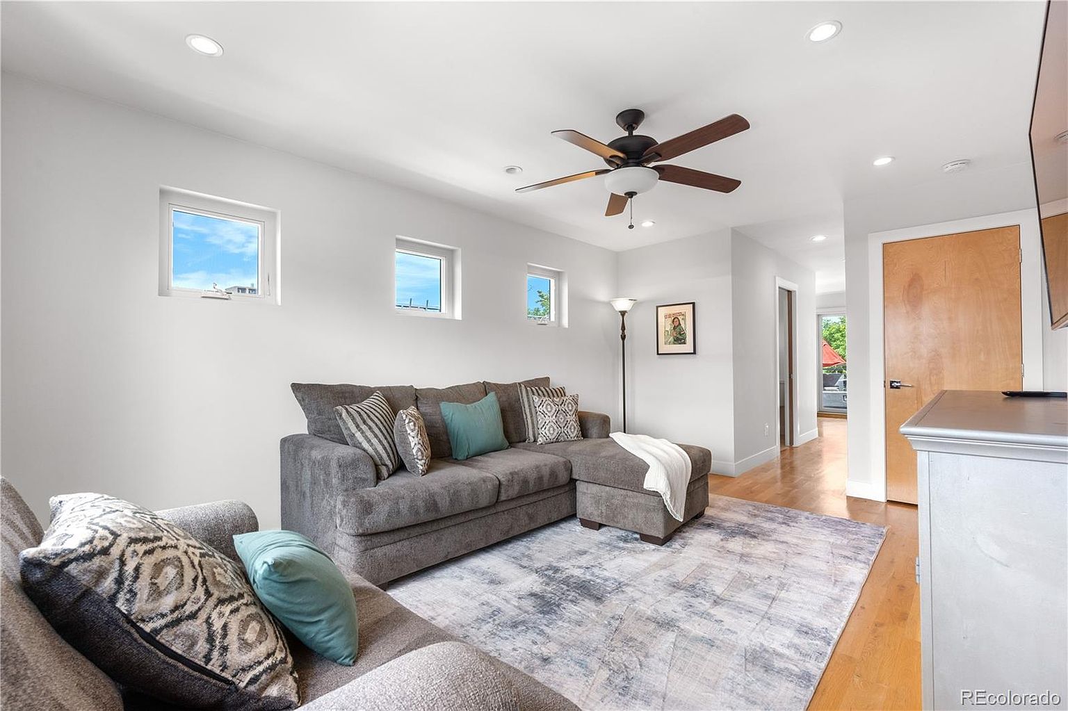 This is an interior shot of a living room featuring a gray sectional sofa with decorative pillows, a patterned area rug, and a ceiling fan. The room has white walls and trim, with natural light coming in through several windows. The overall impression is a comfortable and inviting living space.