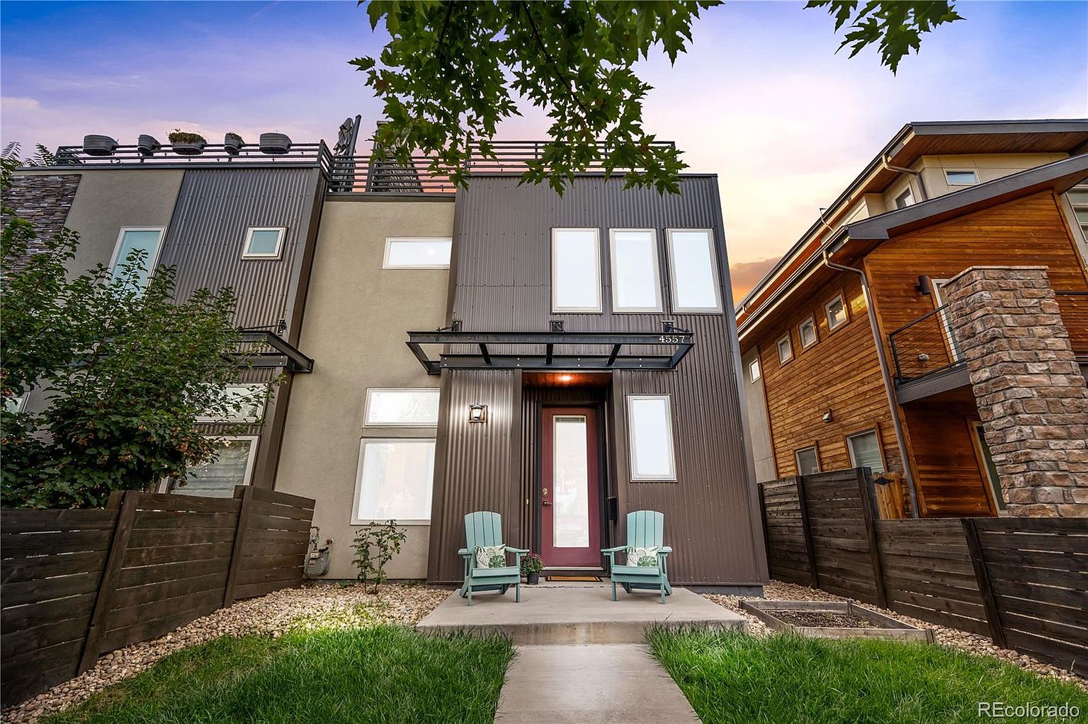 This is a front view of a modern two-story home with a unique architectural design. The exterior features a combination of brown corrugated metal and stucco, with large windows providing ample natural light. A small porch with two light blue chairs and a well-maintained lawn enhance the curb appeal, creating an inviting and contemporary aesthetic.