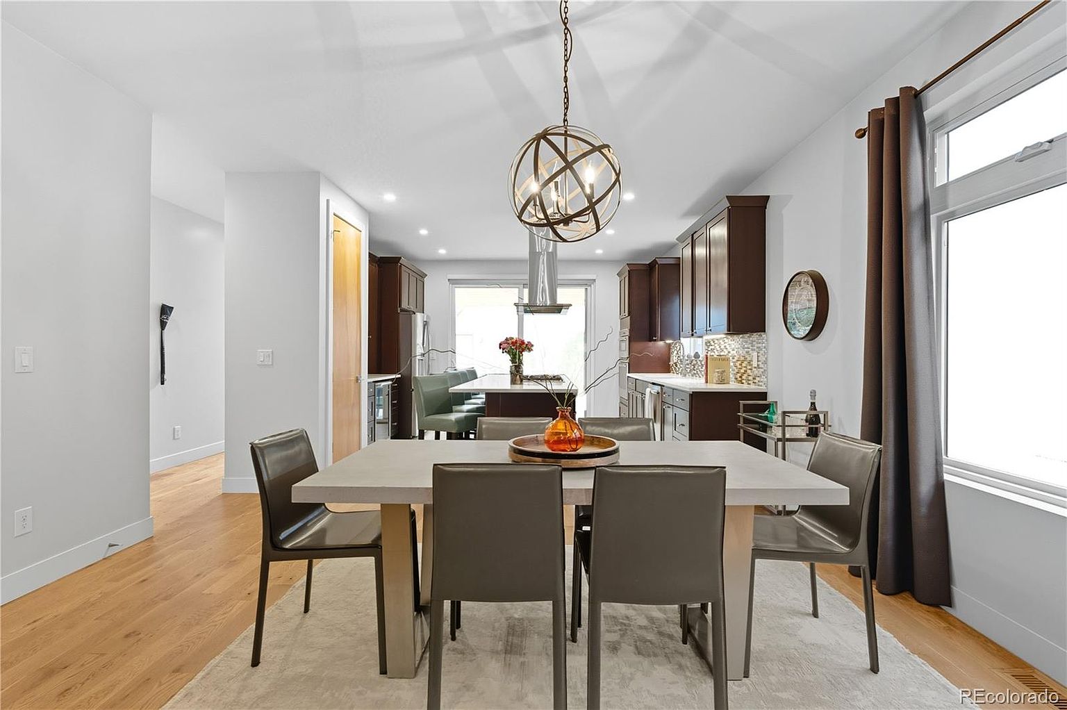 This interior shot showcases a modern dining room with a large rectangular table and six chairs set on a neutral rug. A unique spherical chandelier hangs above the table, and the open floor plan allows a view into the kitchen with dark wood cabinetry and stainless steel appliances. Natural light streams in from a window with brown curtains, creating a bright and inviting atmosphere.