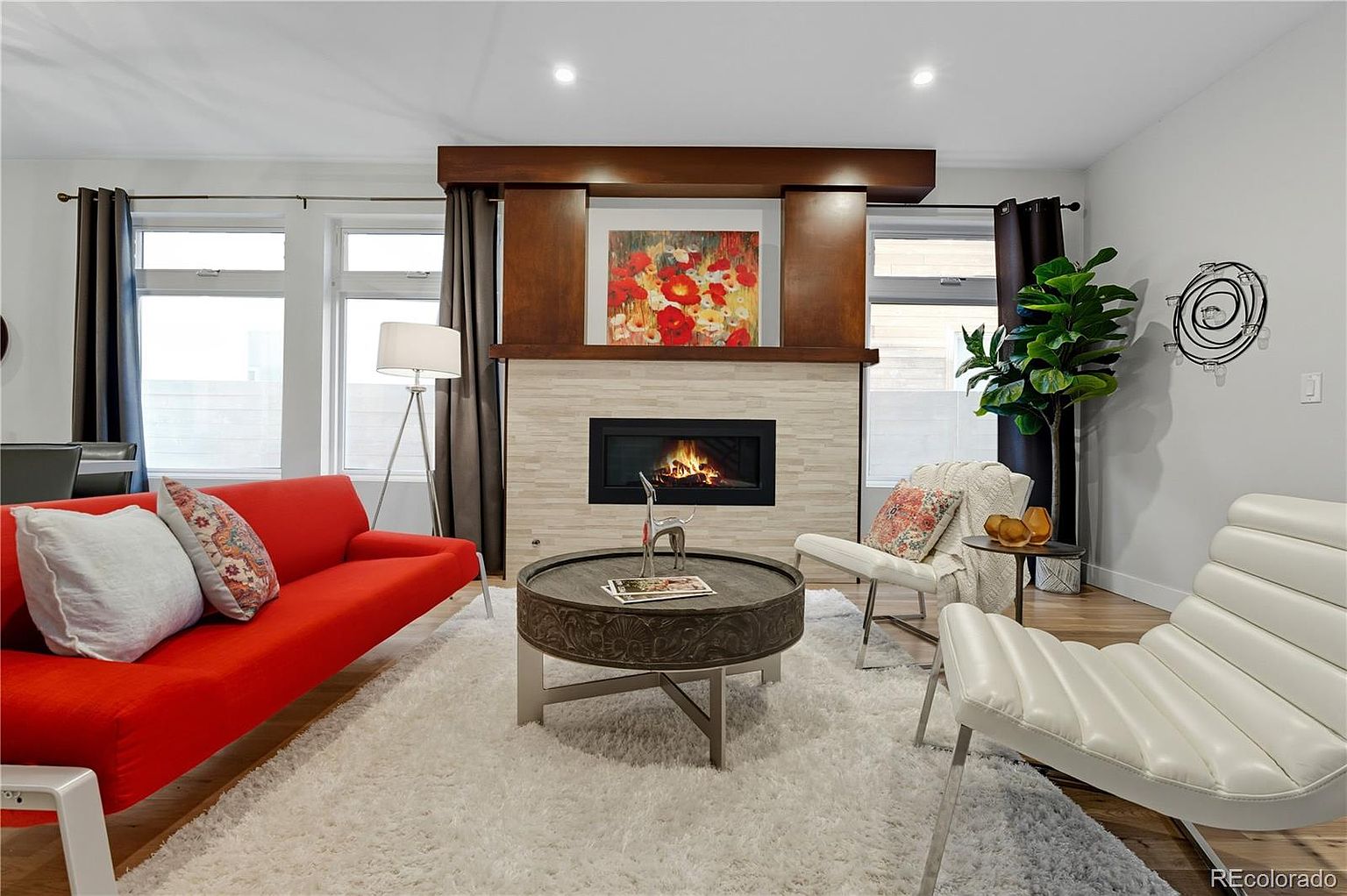 This is an interior shot of a living room featuring a modern fireplace with a wooden mantel and a stone surround. A bright red sofa sits to the left, complemented by white chairs and a unique round coffee table in the center. The room is well-lit with natural light from the windows and features a neutral color palette with pops of color from the artwork and furniture, creating a cozy and stylish atmosphere.