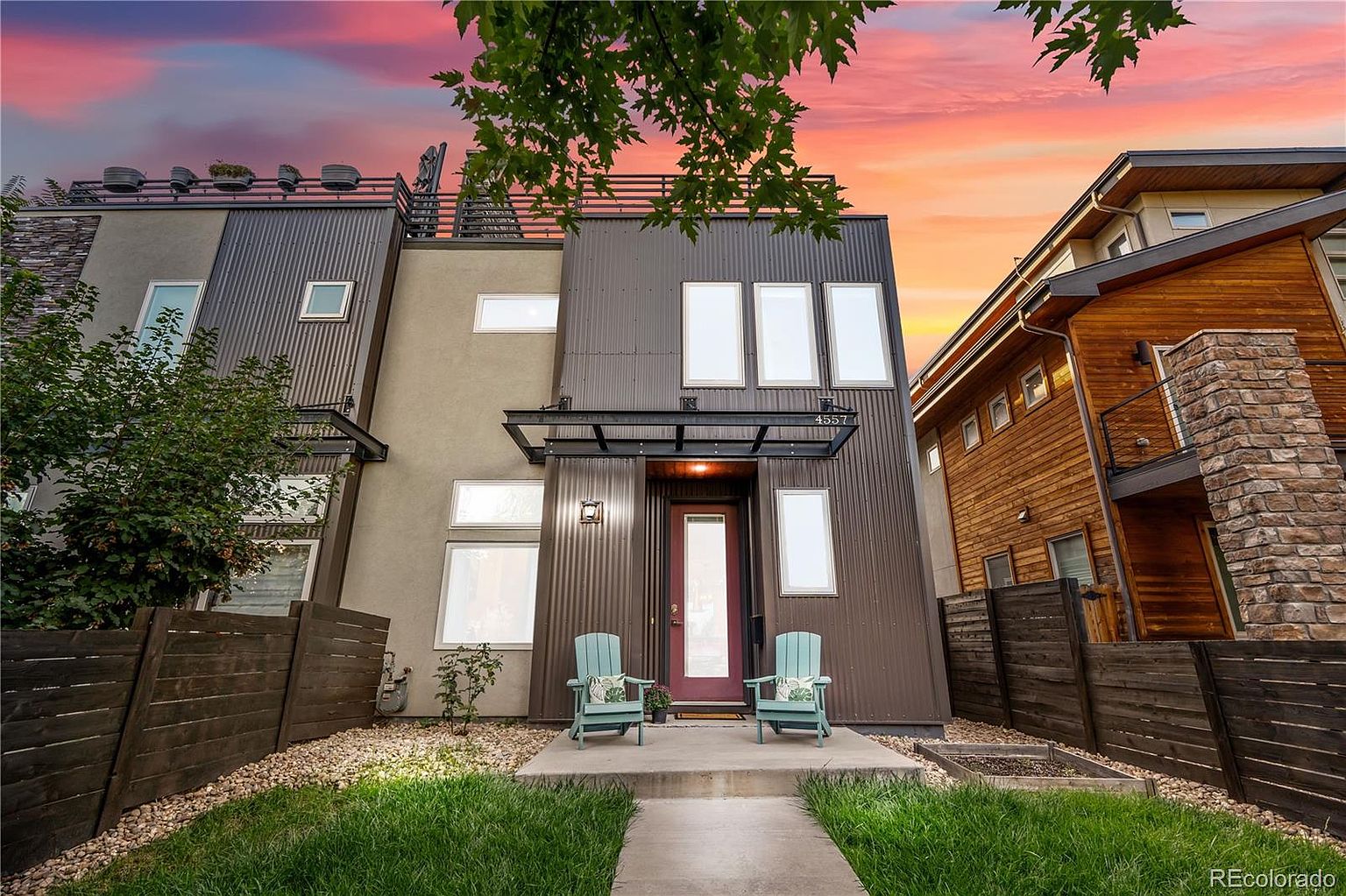The image showcases the front exterior of a modern townhouse with a striking architectural design. The facade features a combination of corrugated metal and stucco, complemented by large windows and a covered entryway with two teal chairs. A well-maintained lawn and landscaping add to the property's curb appeal, creating a welcoming and contemporary aesthetic.