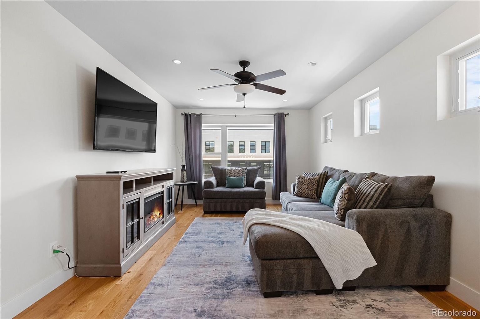 This is a bright and inviting living room featuring a large gray sectional sofa with decorative pillows, a matching armchair, and a stylish area rug. A modern entertainment center with a fireplace sits beneath a wall-mounted television. Natural light floods the room through a large window, creating a warm and comfortable atmosphere.