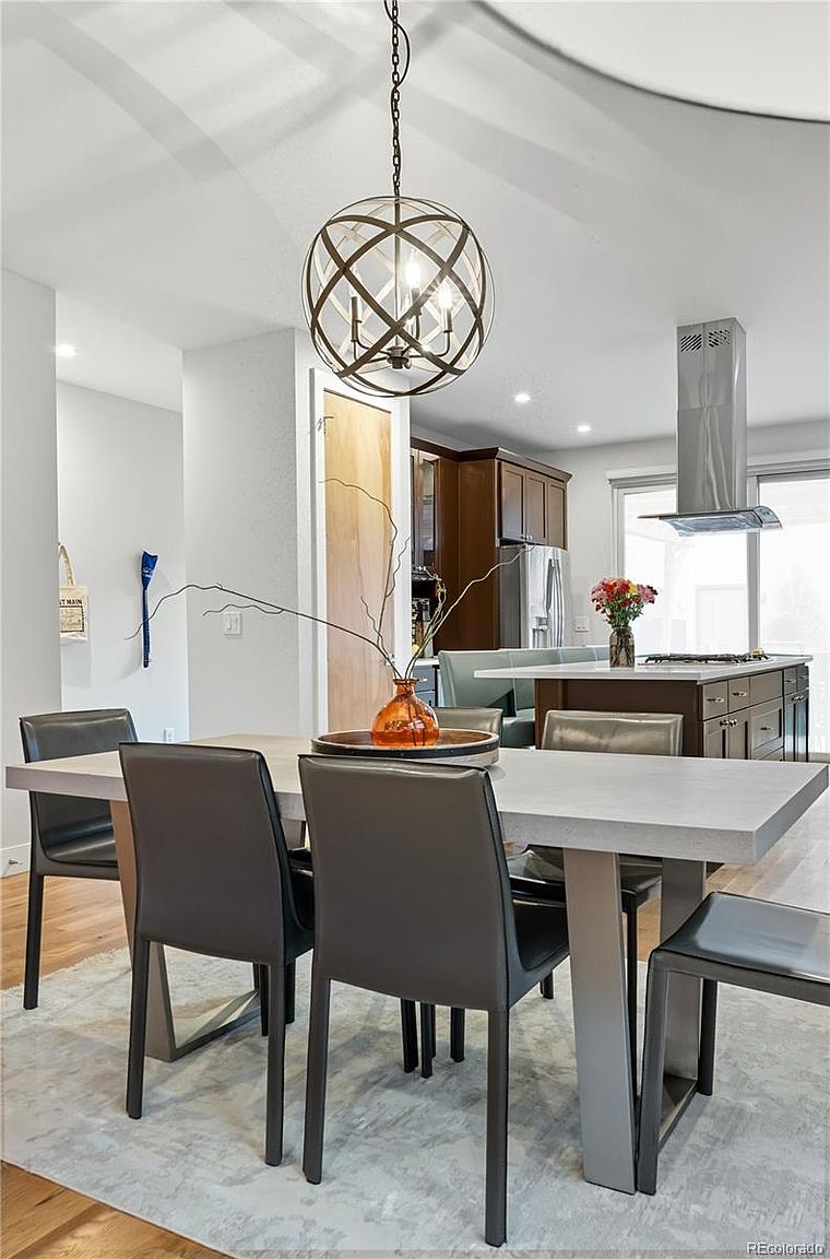 This interior shot showcases a dining room with a modern aesthetic. A rectangular dining table with a light-colored top and unique leg design is surrounded by six dark upholstered chairs. An eye-catching spherical chandelier hangs above the table, and in the background, a glimpse of the kitchen area with stainless steel appliances and dark wood cabinetry can be seen.