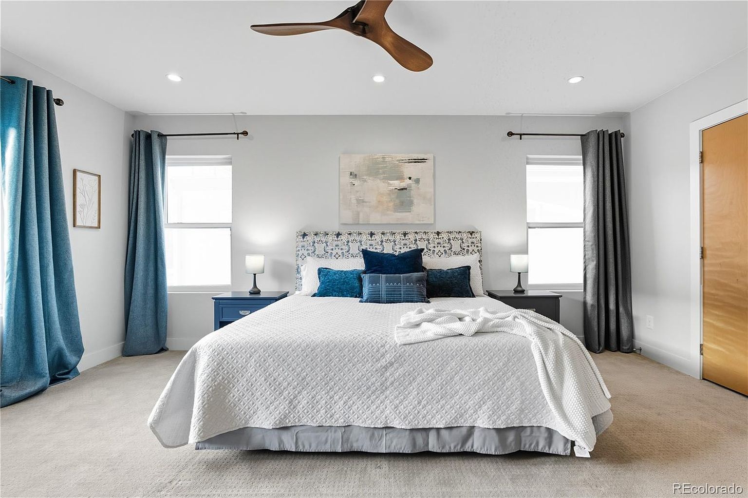 This is a well-lit primary bedroom featuring a bed with a patterned headboard, blue accent pillows, and a white comforter. The room has a neutral color palette with blue and gray curtains flanking windows on either side of the bed. A ceiling fan and recessed lighting provide illumination, and matching nightstands with lamps sit on either side of the bed.