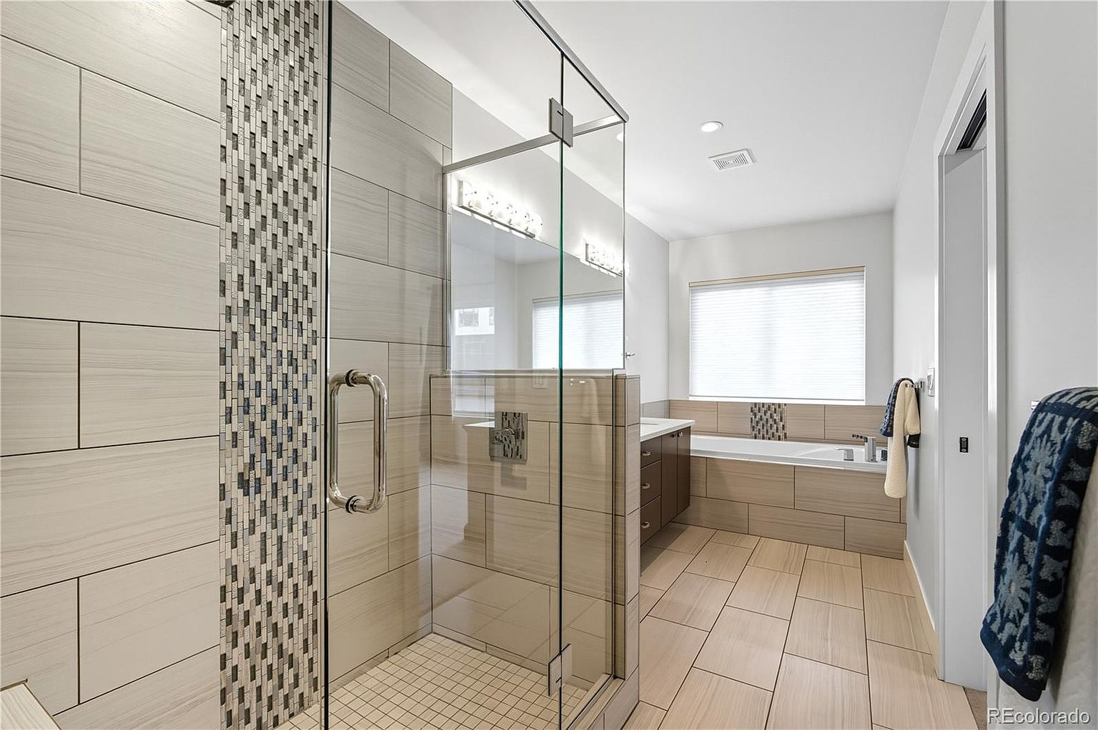 This is a well-lit primary bathroom featuring a glass-enclosed shower with decorative tile accents and a modern shower door. The bathroom includes a built-in bathtub with matching tile surround and a vanity with drawers. The flooring consists of large rectangular tiles, and the overall design is contemporary and clean.