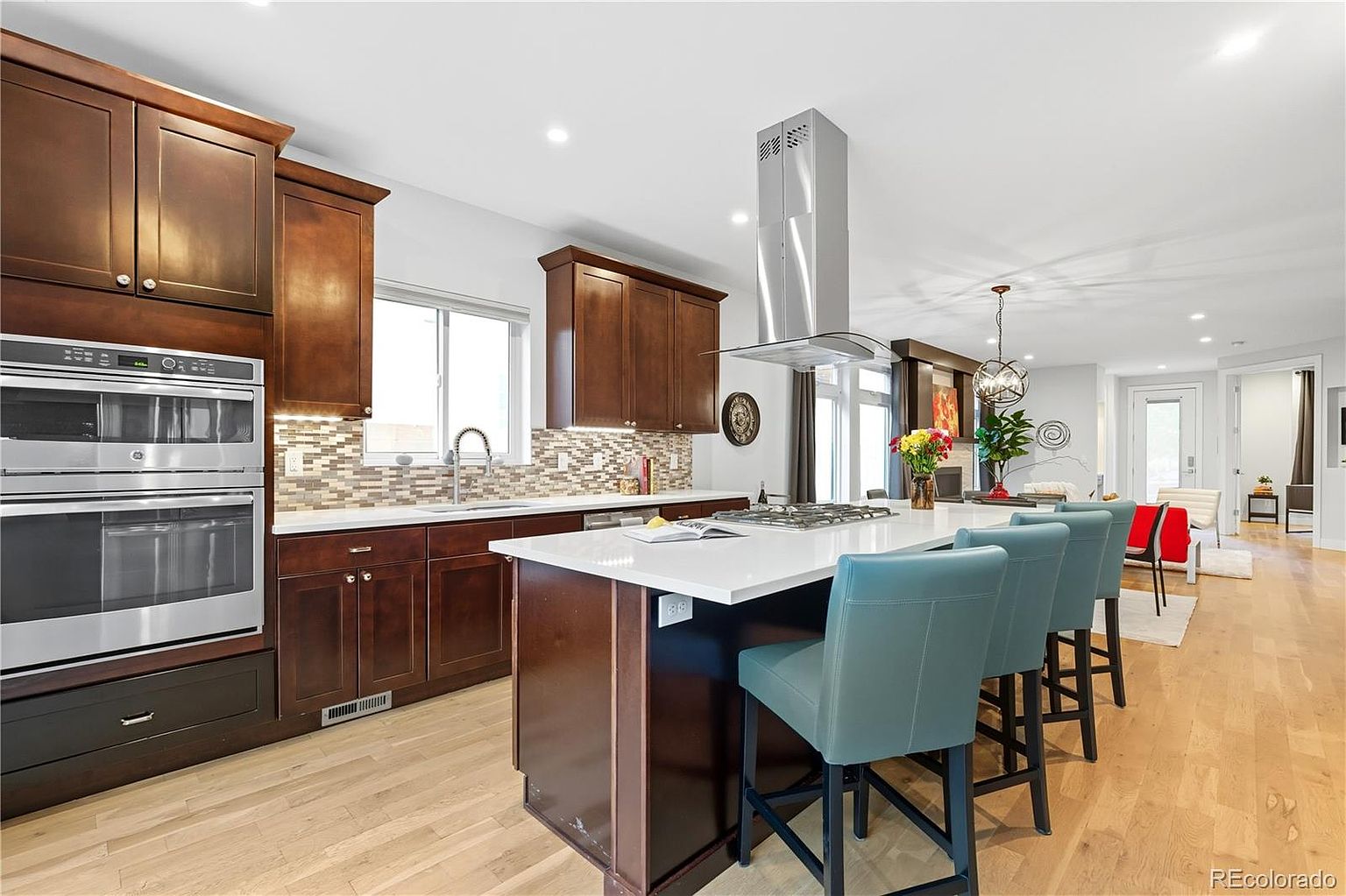 This is a well-lit kitchen featuring dark wood cabinetry, stainless steel appliances, and a white countertop island with seating. The kitchen has a modern design with a mosaic backsplash and an open layout that flows into the living area. The space is clean and inviting, perfect for cooking and entertaining.