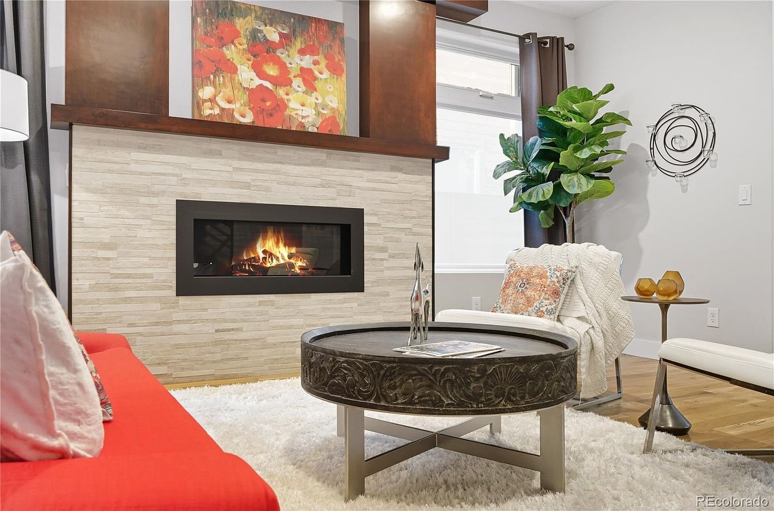 This is an interior shot of a living room featuring a modern fireplace with a stone surround and a dark wood mantel. The room is decorated with a large floral painting above the fireplace, a round coffee table, and a comfortable armchair with a throw blanket. The overall impression is cozy and stylish, perfect for relaxing or entertaining.