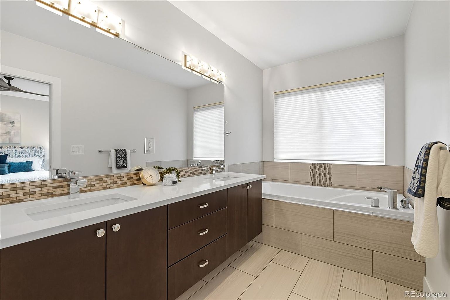 This is a well-lit primary bathroom featuring a double vanity with dark wood cabinets, white countertops, and modern fixtures. A large mirror spans the length of the vanity, reflecting the natural light from a window above the bathtub. The bathtub is surrounded by neutral-toned tiles, creating a spa-like atmosphere.