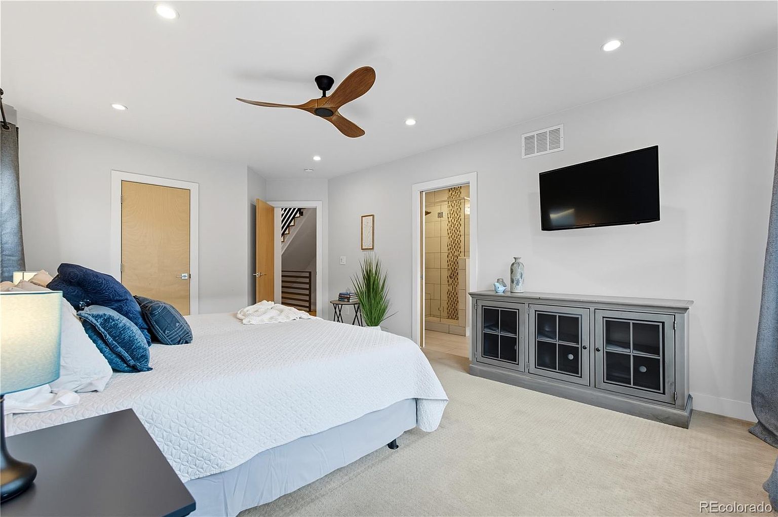 This is a primary bedroom featuring a king-size bed with white bedding and blue pillows. The room has light gray walls, a ceiling fan, and a mounted television above a gray cabinet. There are multiple doorways leading to other areas, including one to a bathroom with a tiled shower.