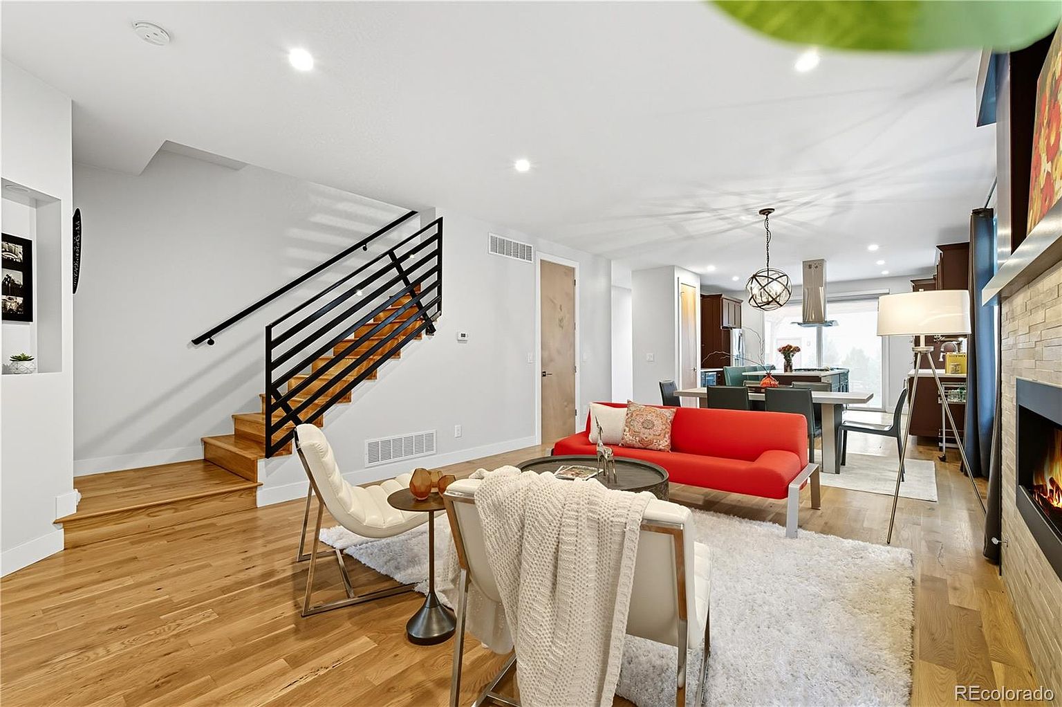 This is an interior shot of a modern living room featuring hardwood floors, a red sofa, and a staircase with black railings. The room is well-lit with recessed lighting and natural light, creating a bright and inviting atmosphere. The open floor plan connects the living room to the dining area and kitchen, enhancing the sense of space.