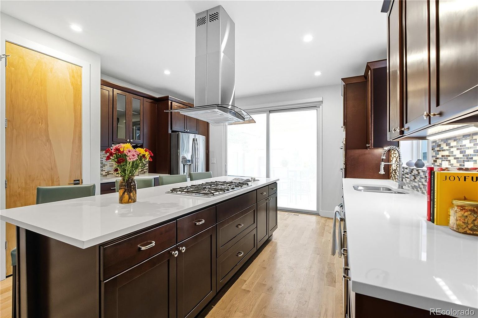 This is a well-lit kitchen featuring dark wood cabinetry and a large island with a white countertop. Stainless steel appliances and a modern range hood add a contemporary touch. The kitchen also has a sliding glass door that provides natural light and access to the outside.