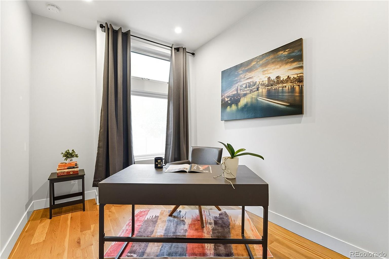 This is an interior shot of a home office. The room features a modern desk with a plant and a mug on top, a colorful rug underneath, and a piece of art hanging on the wall. A window with gray curtains provides natural light, and a small side table with books and a plant sits to the left.