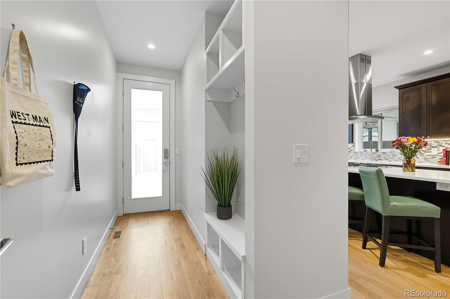 This is an interior shot of a bright and modern hallway. The hallway features light wood flooring, white walls, and a built-in storage unit with shelves and hooks. A front door with glass panels is visible at the end of the hallway, and a glimpse of the kitchen can be seen on the right.