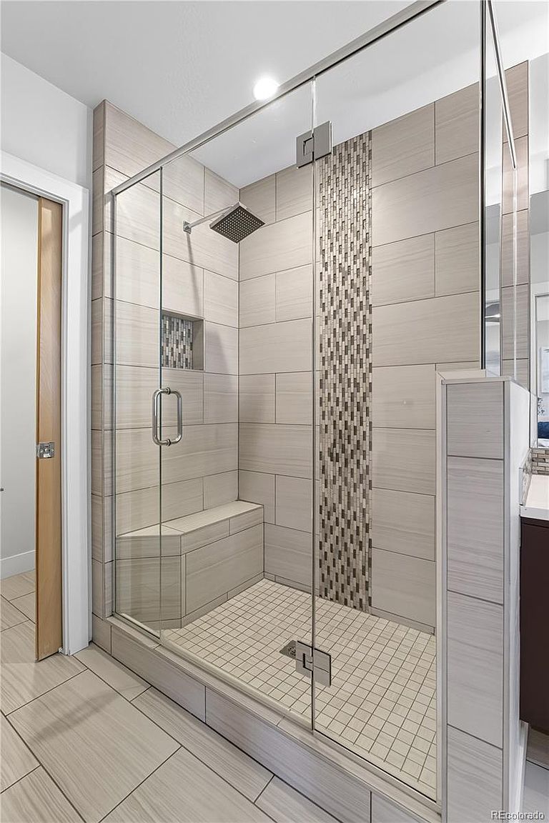 This is a well-lit primary bathroom featuring a modern glass-enclosed shower with a built-in bench and a decorative mosaic tile accent. The shower has a square rain shower head and a niche for toiletries. The floor and wall tiles are a light neutral color, creating a clean and spa-like atmosphere.