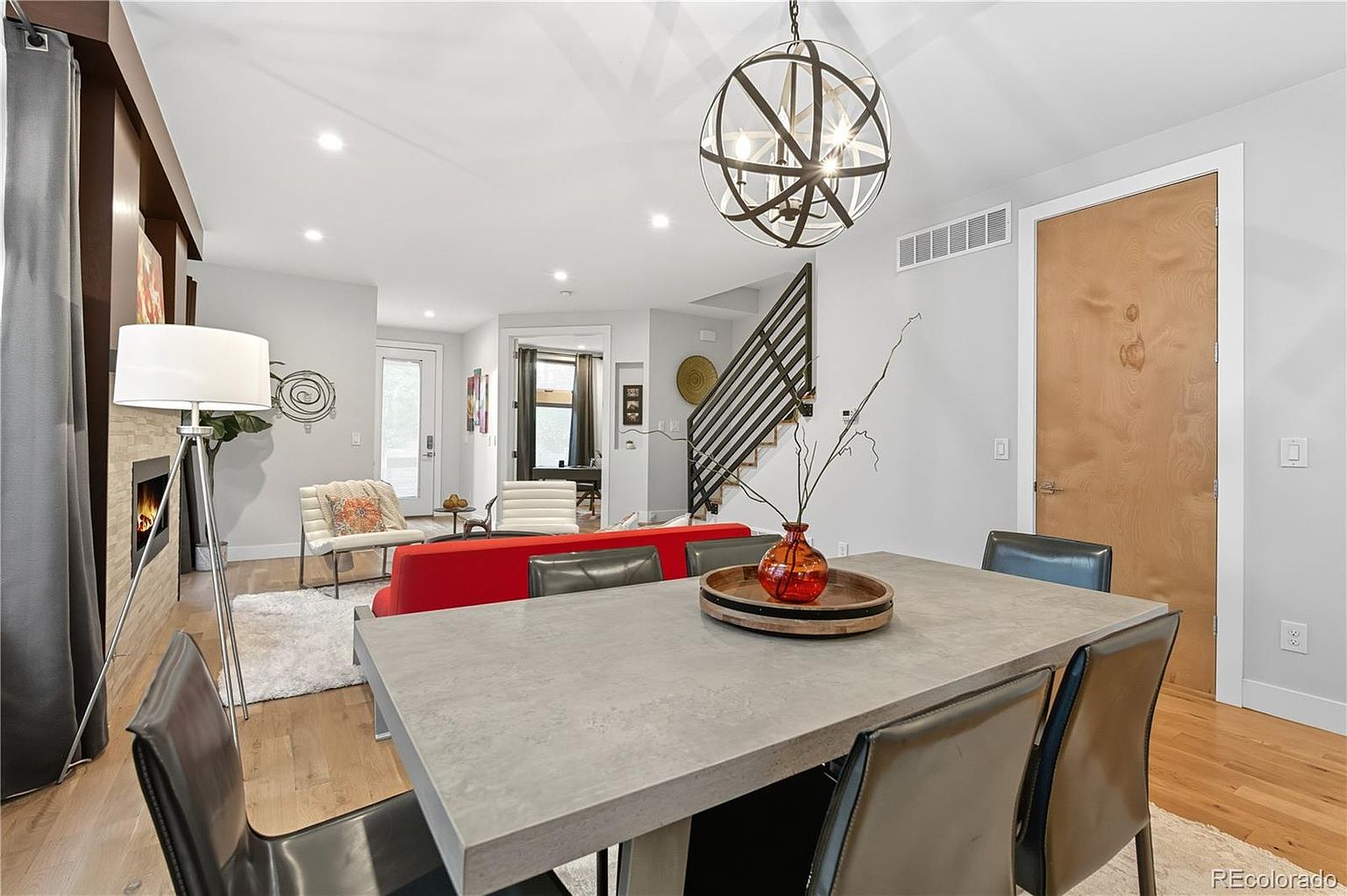 This interior shot showcases a modern dining room with a large concrete table surrounded by leather chairs. An eye-catching spherical chandelier hangs above the table, and a red sofa adds a pop of color in the background. The room flows seamlessly into the living area, creating an open and inviting space.