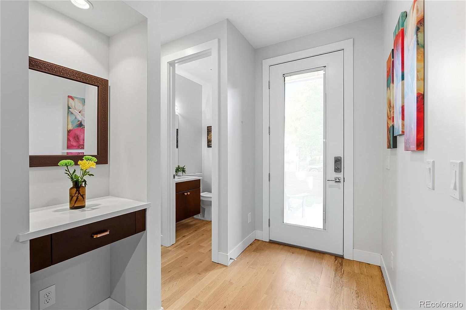 This is an interior shot of a hallway featuring light hardwood floors and white walls. A console table with a mirror above it sits to the left, while a doorway leads to a bathroom. A white front door with a large glass panel is visible on the right, with modern artwork hanging nearby, creating a bright and welcoming entryway.