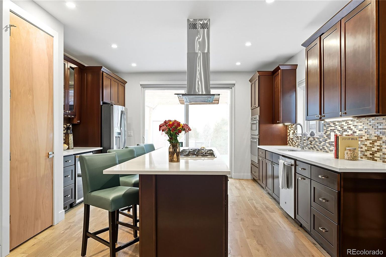 This is a well-lit kitchen featuring dark wood cabinetry and stainless steel appliances. A large island with a white countertop and seating provides a central gathering space, while a modern range hood hangs above the cooktop. The hardwood floors and mosaic backsplash add warmth and texture to the space, creating an inviting atmosphere.