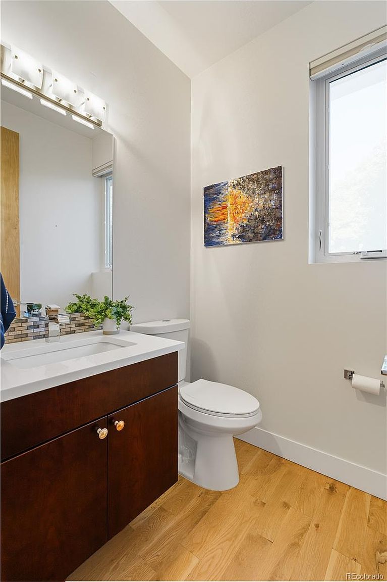 This is a well-lit guest bathroom featuring a modern vanity with a dark wood cabinet and a white countertop. A toilet is positioned next to the vanity, and an abstract painting hangs on the wall above it. The light wood flooring adds warmth to the space, creating a clean and inviting atmosphere.