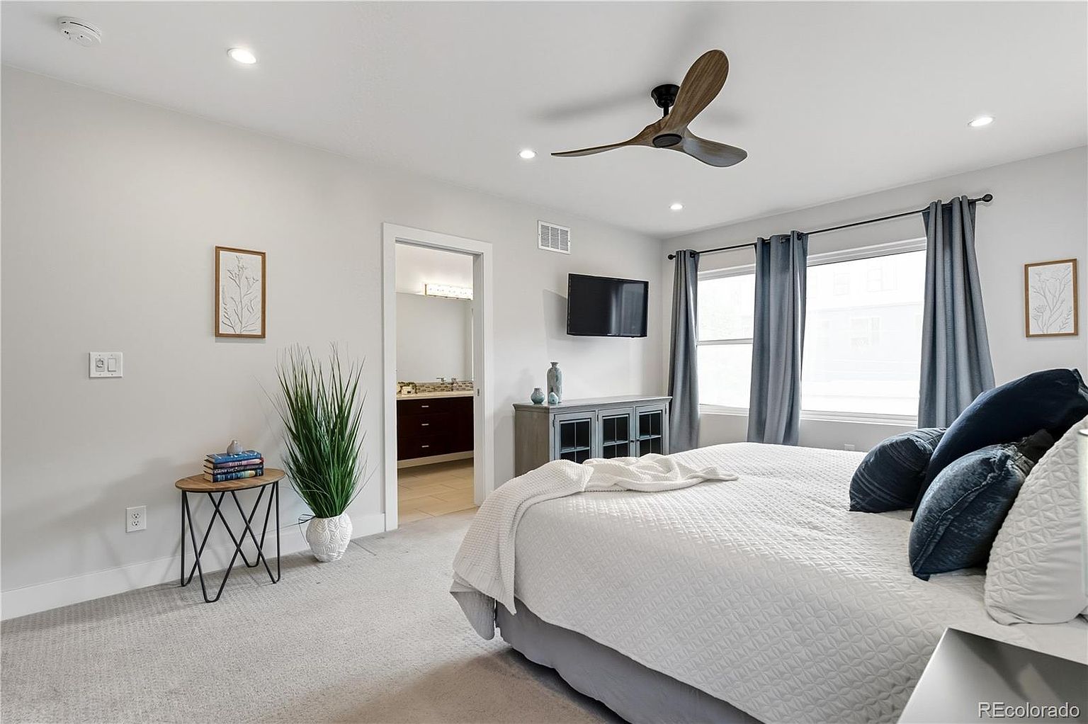 This is a bright and airy primary bedroom featuring a large bed with white bedding and blue decorative pillows. The room has a modern ceiling fan, a wall-mounted television, and a console table. Natural light streams in through the windows, which are framed by blue curtains, and the walls are painted in a neutral tone, creating a serene and inviting atmosphere.