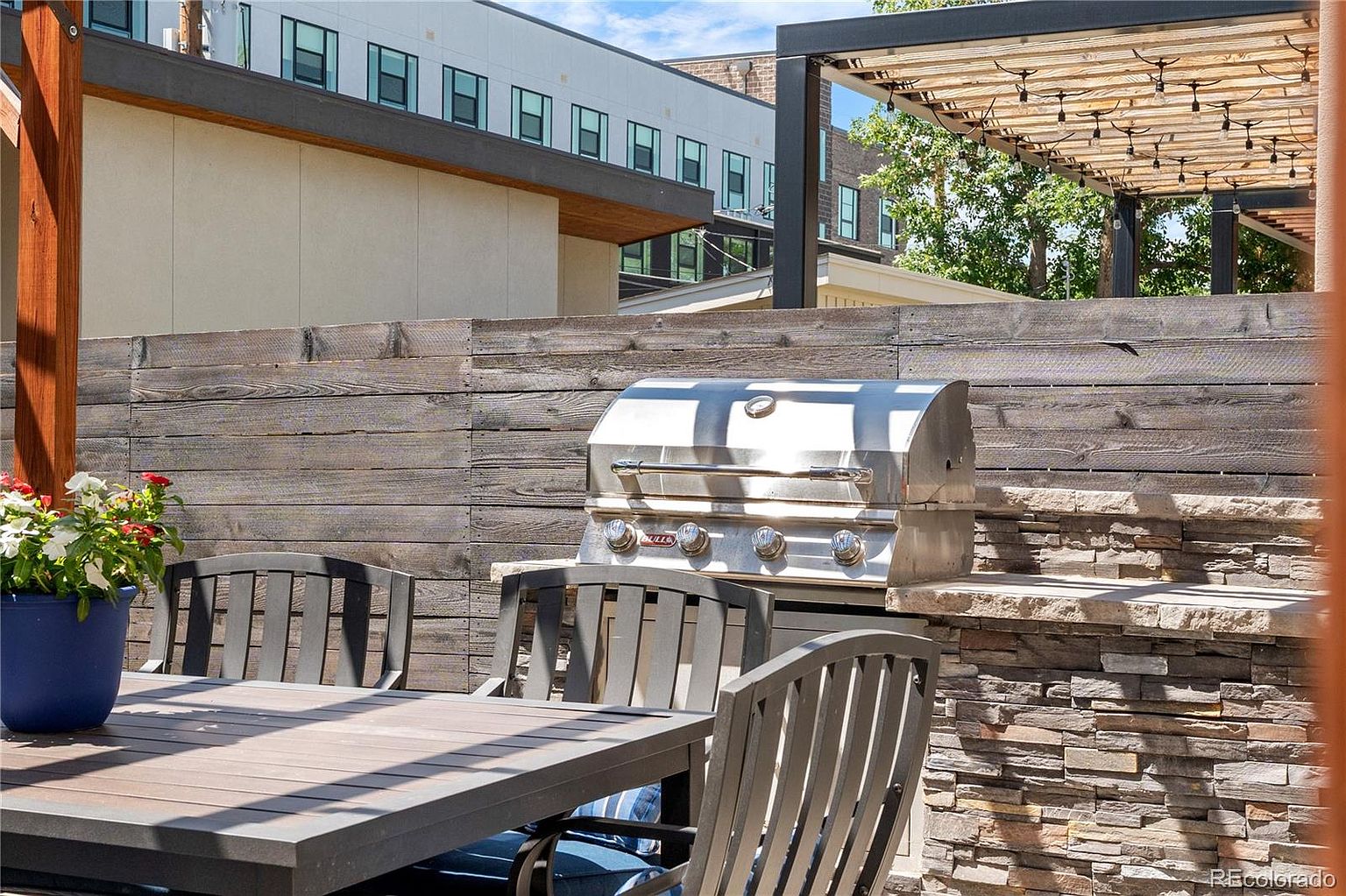 This image showcases an inviting outdoor patio area, perfect for entertaining. A modern stainless steel grill sits against a stone-clad counter, complemented by a wooden privacy fence. A dark-colored dining table with matching chairs is positioned nearby, suggesting a comfortable space for outdoor meals.