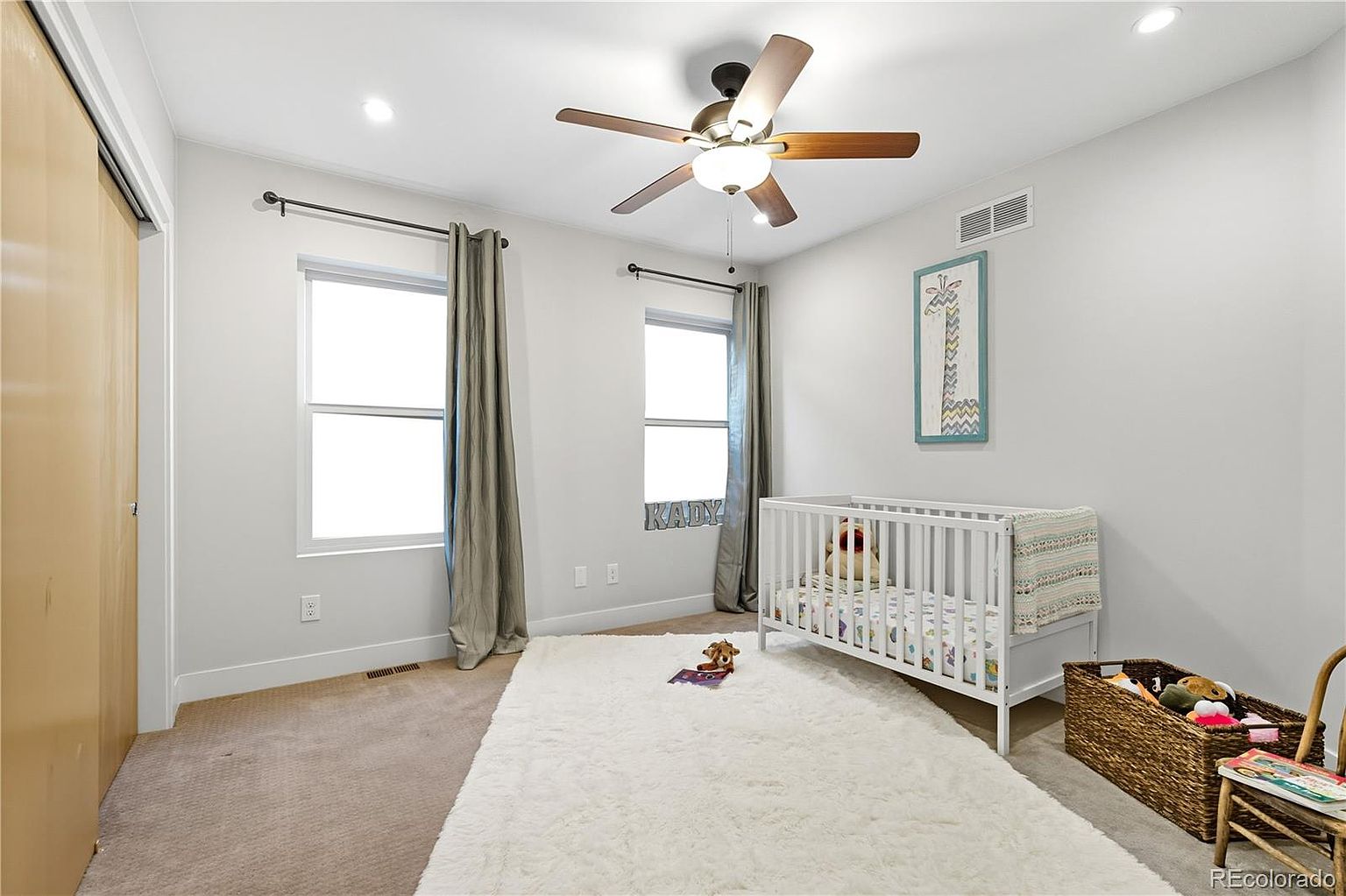 This is a cozy bedroom featuring a white crib with a patterned blanket and stuffed animals, suggesting it's a nursery. The room has two windows with gray curtains, a ceiling fan, and a fluffy white rug that adds warmth. A woven basket with toys and a small chair are also visible, contributing to the room's inviting and child-friendly atmosphere.