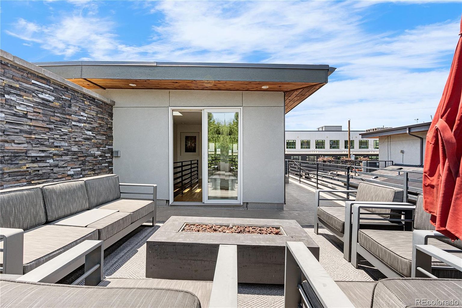 This image showcases a modern outdoor patio or balcony area. It features stylish gray outdoor furniture arranged around a concrete fire pit, creating a cozy and inviting atmosphere. The space is enhanced by a stone accent wall and a sliding glass door leading indoors, offering a seamless transition between indoor and outdoor living.