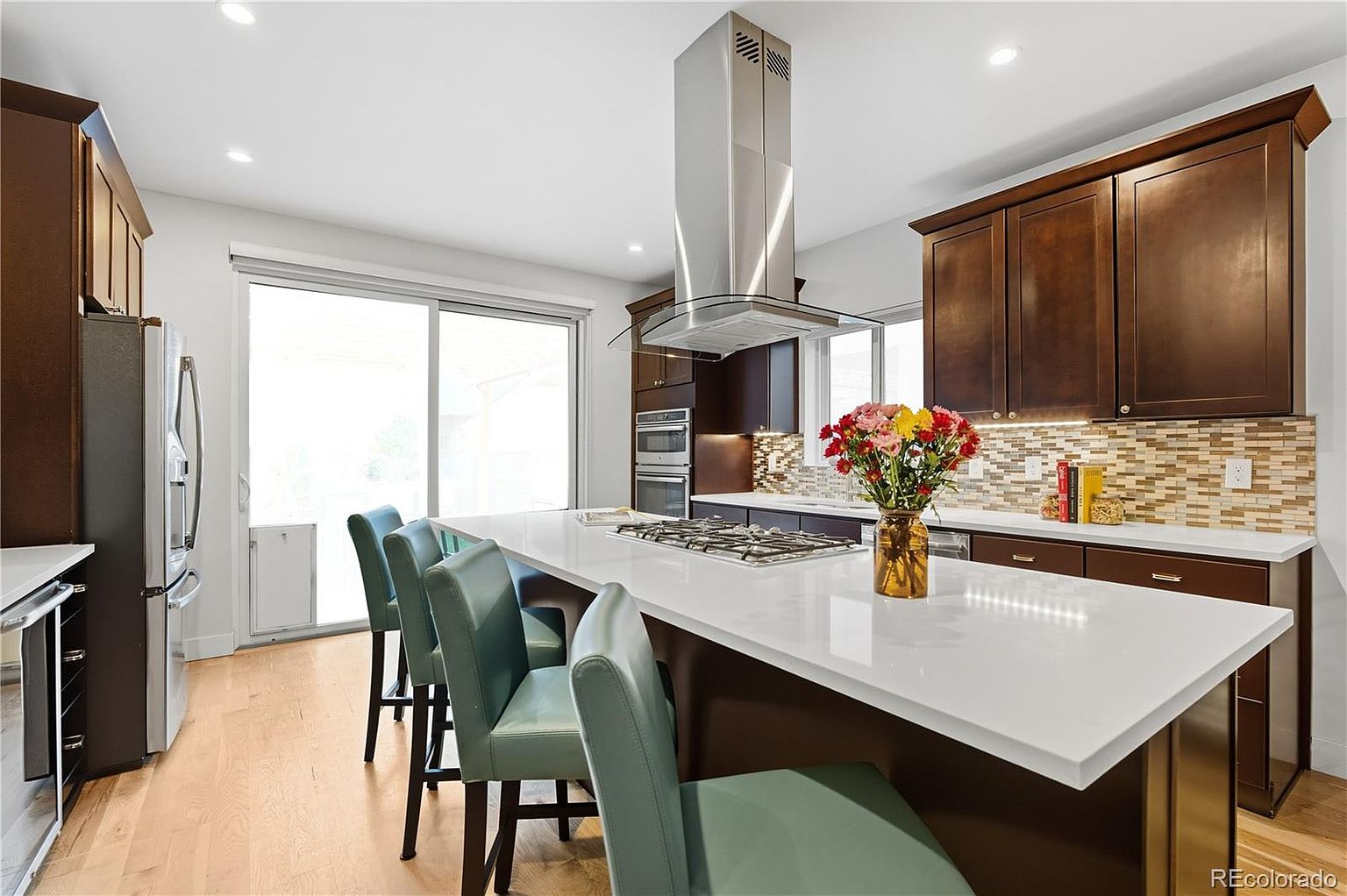 This is a well-lit kitchen featuring a large island with a white countertop and seating for three. The kitchen has dark wood cabinets, stainless steel appliances, and a mosaic backsplash. A bouquet of flowers adds a pop of color to the island, and a sliding glass door provides access to the outside.
