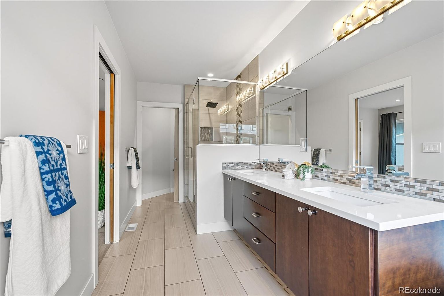 This is a well-lit primary bathroom featuring a double vanity with dark wood cabinets and a white countertop. A large mirror spans the length of the vanity, reflecting the lighting and creating a sense of spaciousness. The bathroom also includes a glass-enclosed shower and neutral-toned tile flooring, contributing to a clean and modern aesthetic.