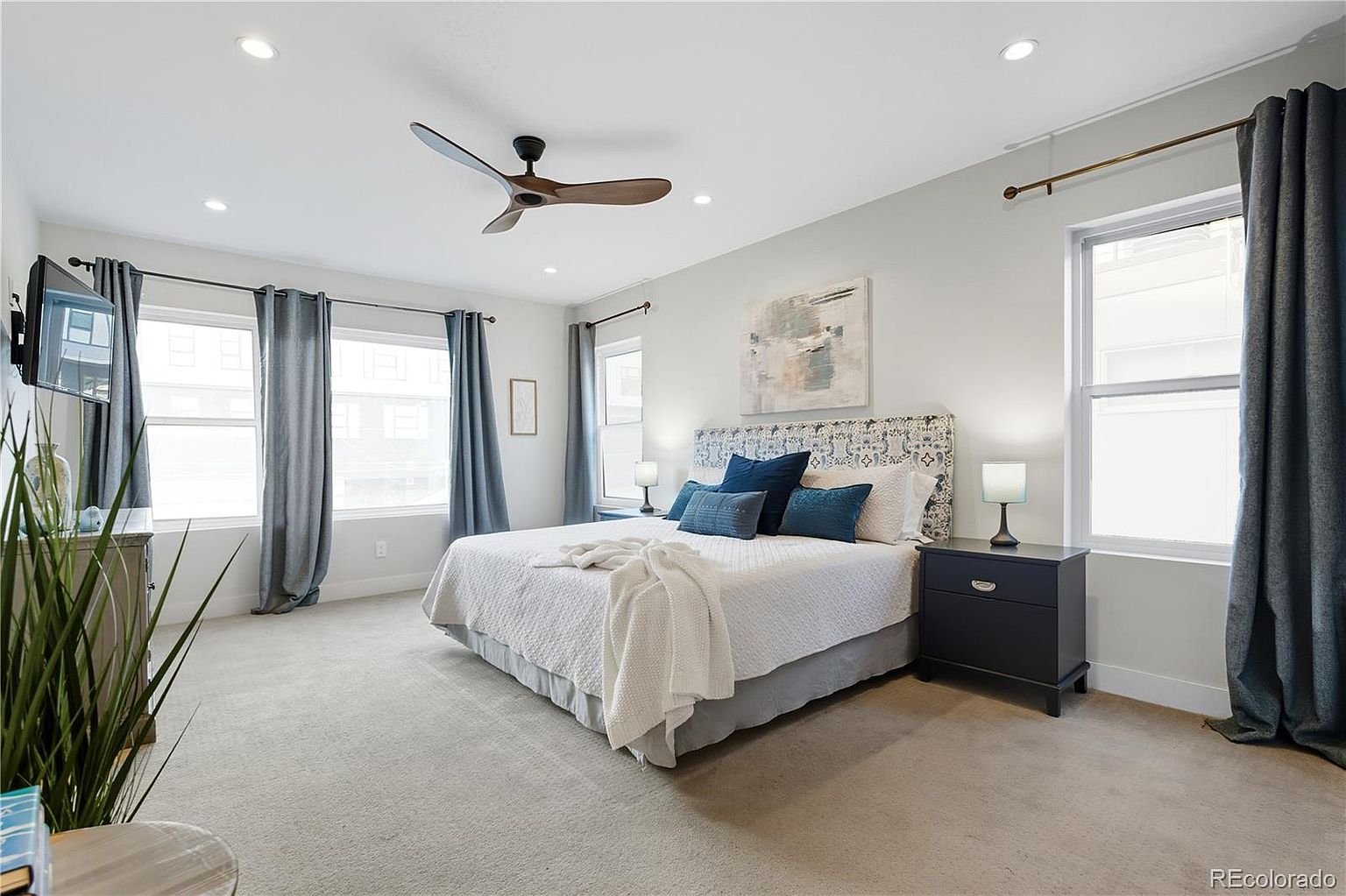 This is a well-lit primary bedroom featuring a bed with a patterned headboard and several blue pillows. The room has a neutral color palette with light gray walls and carpet, complemented by blue curtains framing the windows. A dark-colored nightstand sits beside the bed, and an abstract painting hangs on the wall, creating a serene and inviting atmosphere.