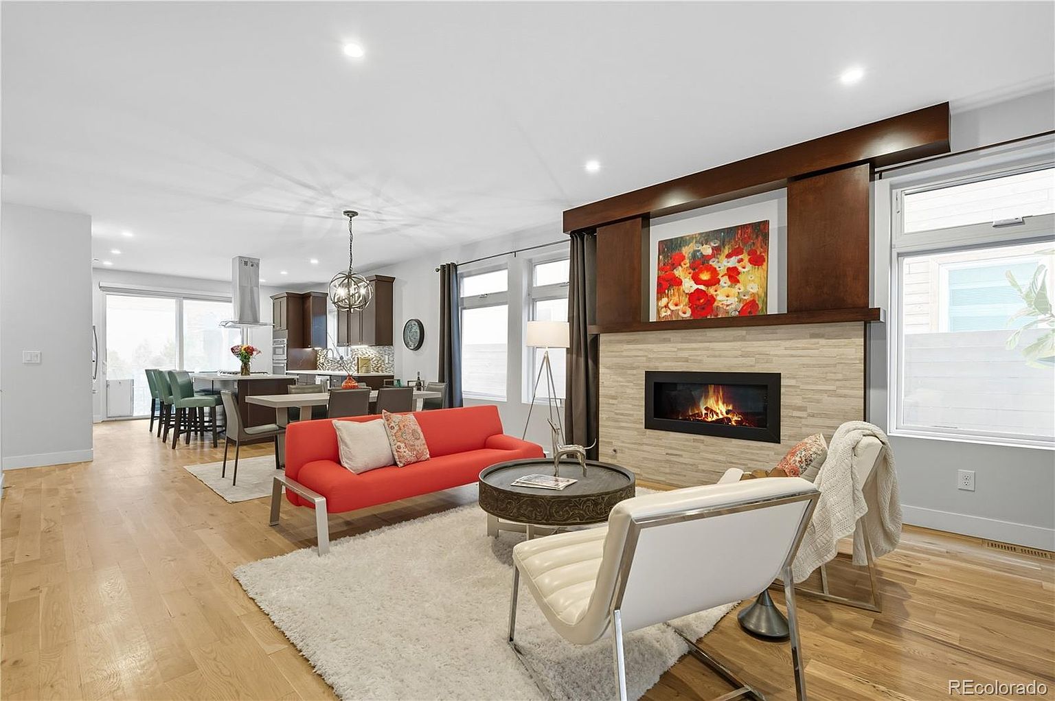 This is an interior shot of a modern living room featuring a fireplace with a stone surround and a dark wood mantel. The room is furnished with a bright orange sofa, a round coffee table, and a white chair, all arranged on a plush white rug. The open floor plan connects to a kitchen area with bar seating, creating a spacious and inviting atmosphere.