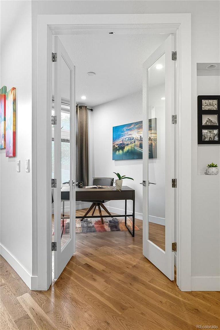This is an interior shot of a home office, viewed through a set of double doors. The office features a modern desk, a comfortable chair, and artwork on the walls. The hardwood flooring adds warmth to the space, and the overall impression is one of a stylish and functional workspace.