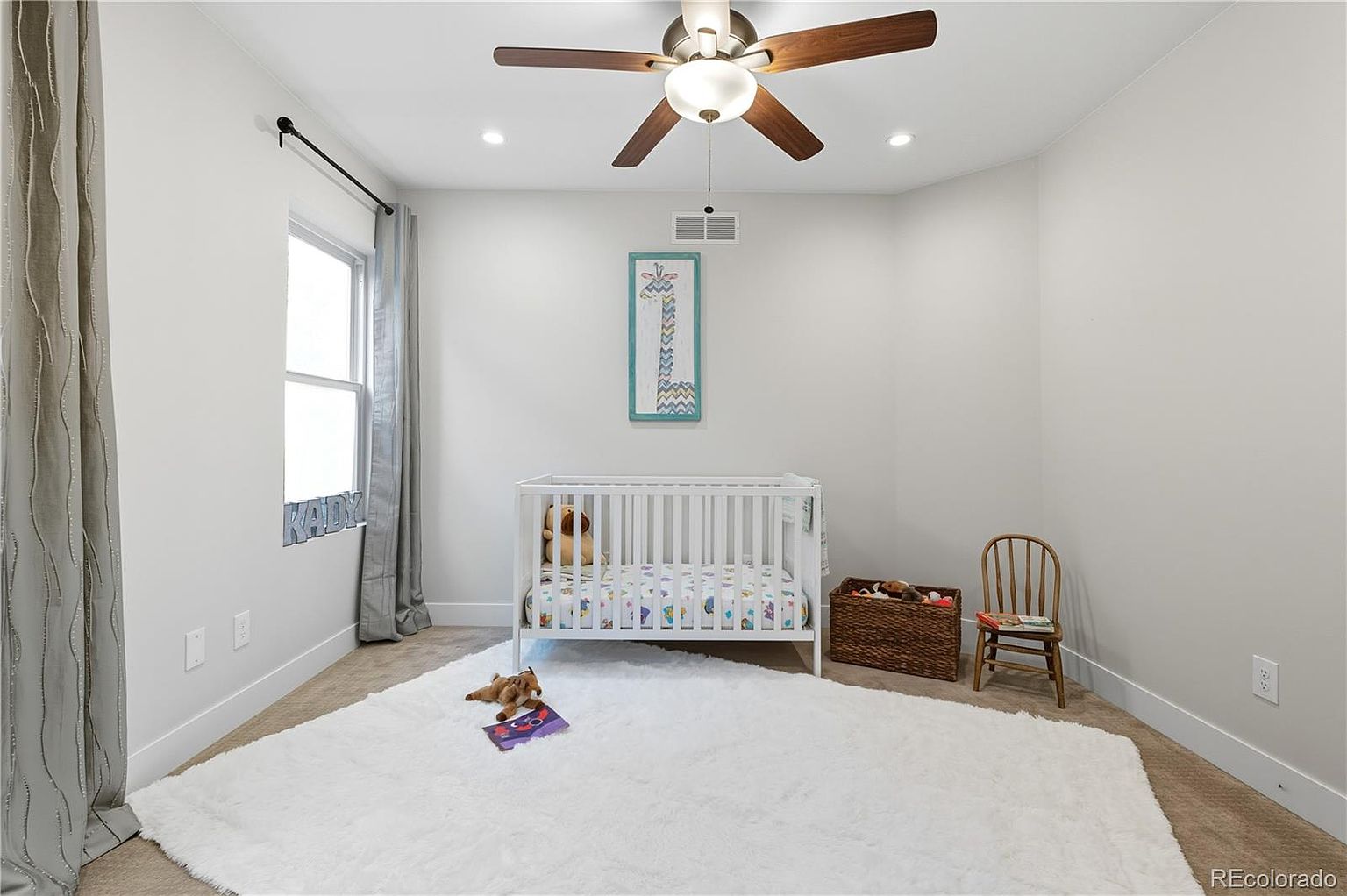 This is a cozy nursery featuring a white crib with colorful bedding, a white fluffy rug, and a wooden chair. The walls are painted in a neutral tone, and a ceiling fan provides air circulation. A window with gray curtains allows natural light to fill the room, creating a warm and inviting atmosphere.