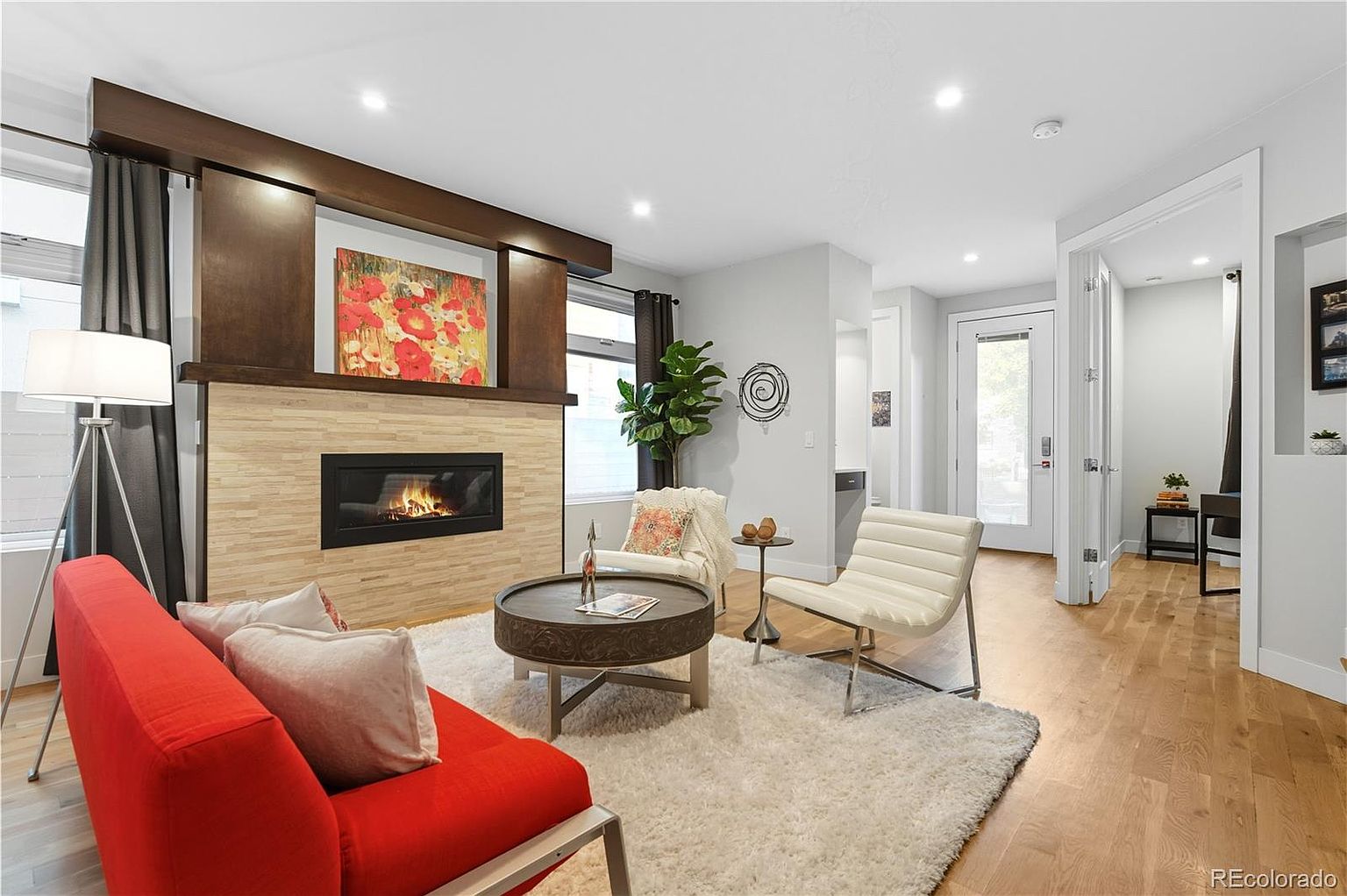 This is an interior shot of a living room featuring a modern fireplace with a wooden mantel and a stone facade. The room is furnished with a red sofa, a round coffee table, and two white chairs, all arranged on a plush white rug. Natural light streams in through a window, complemented by recessed lighting, creating a warm and inviting atmosphere.