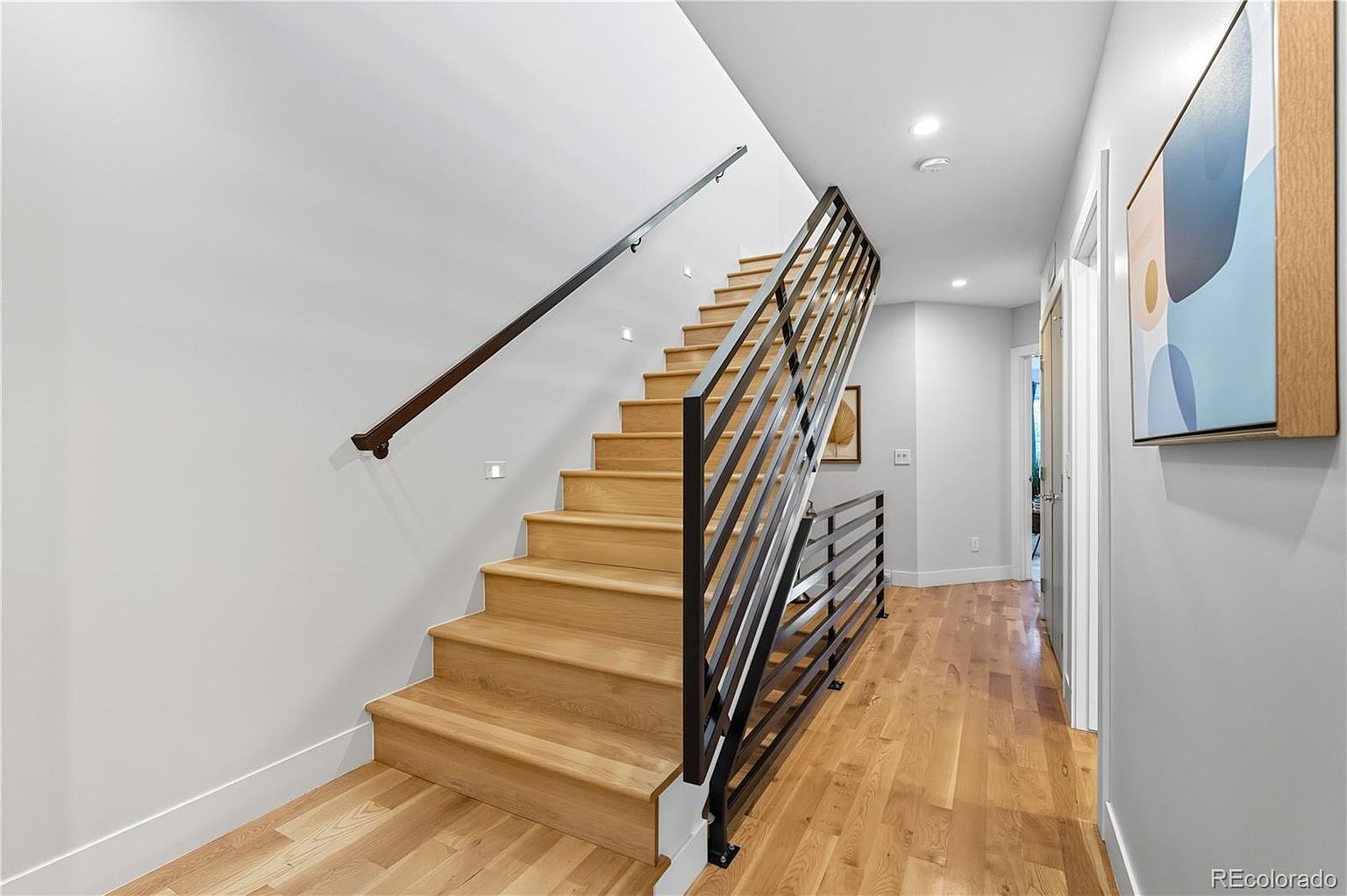This interior shot showcases a modern hallway and staircase. The staircase features light wood steps and a sleek, dark metal railing, contrasting with the light gray walls. The hallway has hardwood flooring and leads to other rooms, with a contemporary art piece adding a touch of sophistication.