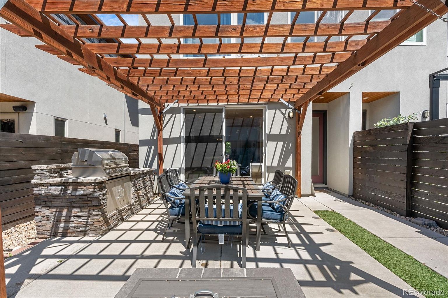 This is an inviting outdoor patio area featuring a wooden pergola that casts interesting shadows on the concrete surface. A dining table with chairs and blue cushions sits beneath the pergola, creating a comfortable space for outdoor meals. A built-in grill area with stone accents adds to the functionality of the space, while a strip of artificial turf provides a touch of greenery.