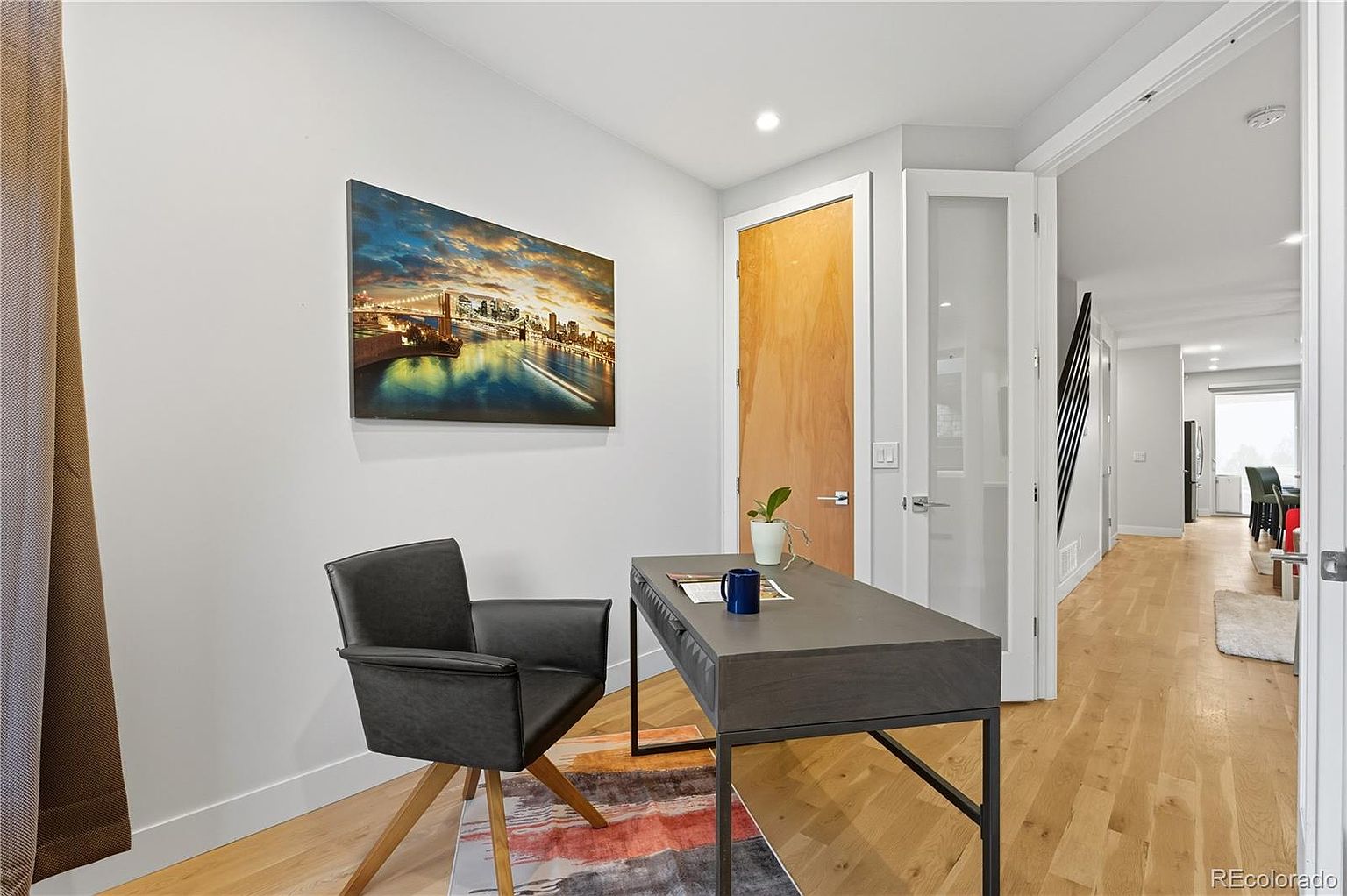 This is an interior shot of a home office. The room features a modern desk with a dark gray finish and black metal legs, paired with a black leather chair with wooden legs. A colorful abstract rug sits beneath the desk, and a cityscape artwork hangs on the wall. The room is well-lit and has a clean, contemporary feel.