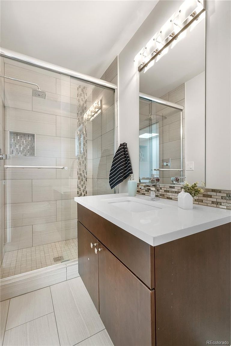This is a well-lit bathroom featuring a modern vanity with a dark wood finish and a white countertop. A large mirror with integrated lighting hangs above the sink, reflecting the adjacent shower with glass doors and tiled walls. The overall impression is clean and contemporary, with a focus on functionality and style.