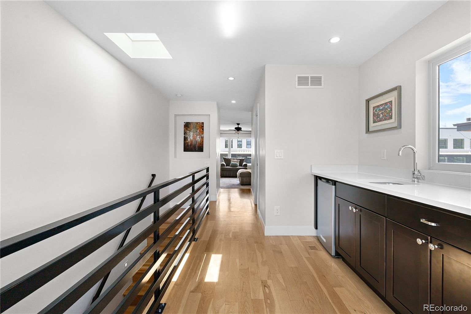 This interior shot showcases a bright hallway with hardwood floors and a modern black railing. A wet bar with dark cabinetry and a white countertop is featured on the right, illuminated by natural light from a window. The hallway leads to a living area, creating a sense of flow and spaciousness.