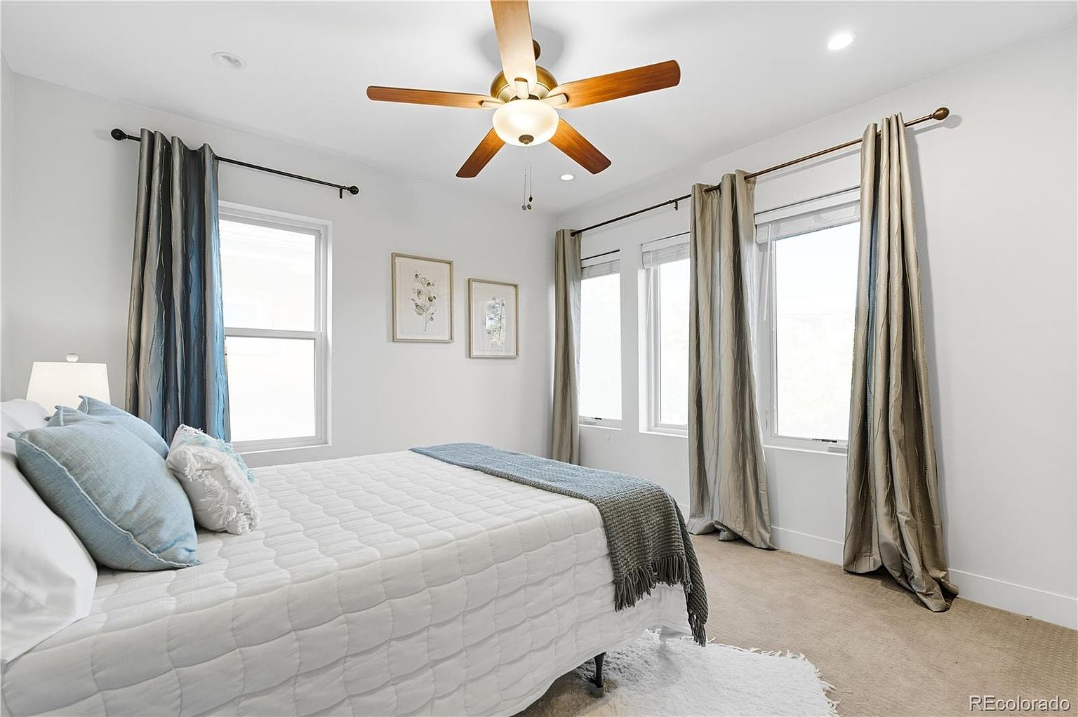 This is a bright and airy primary bedroom featuring a white quilted bed with blue and white pillows, flanked by windows with neutral-toned curtains. The room has a ceiling fan with wooden blades and light fixtures, and artwork hangs on the white walls. The overall impression is clean, comfortable, and inviting.