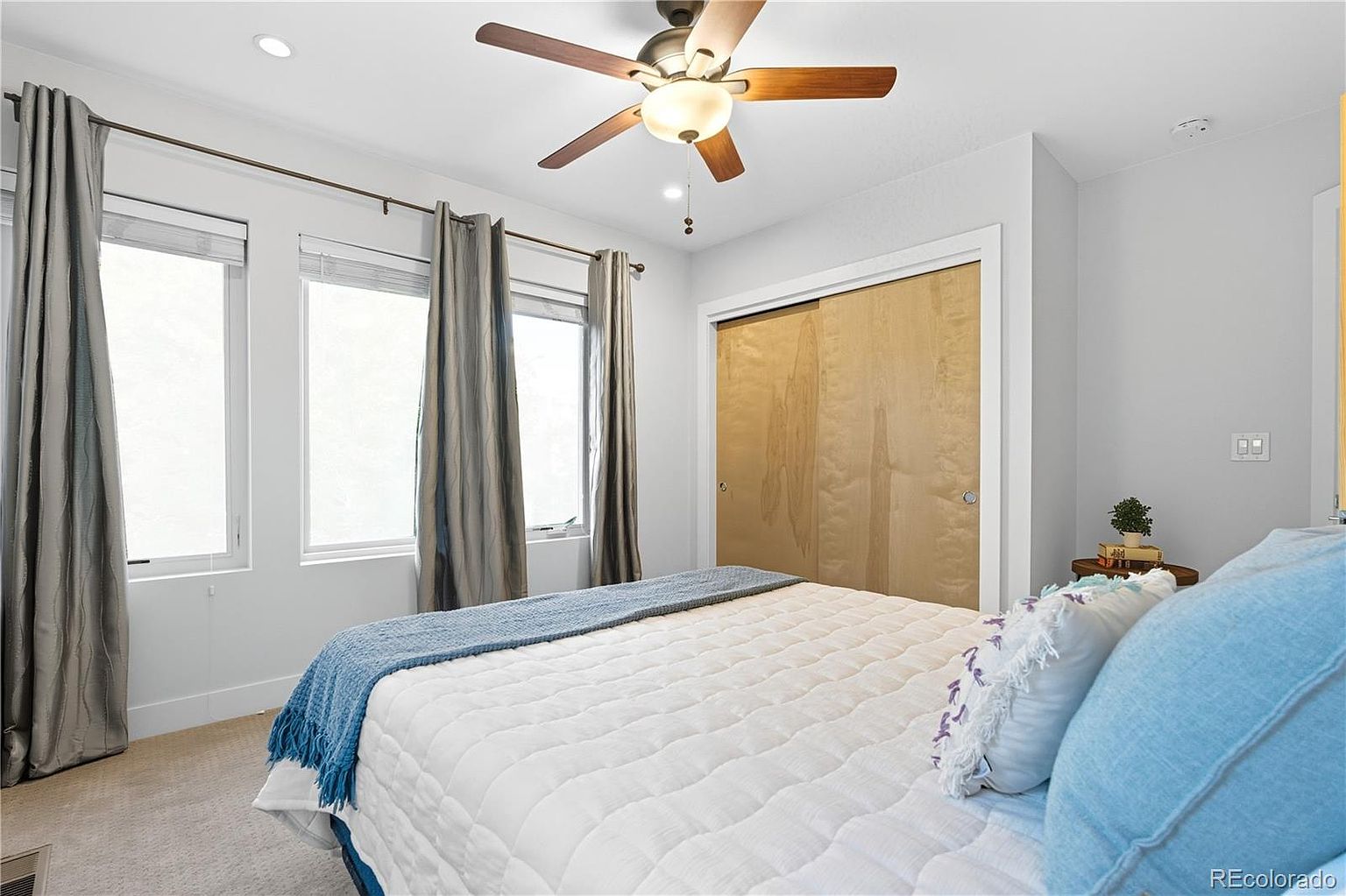 This is a well-lit primary bedroom featuring a bed with a white quilted comforter and blue accents. The room has a neutral color palette with light gray walls, complemented by gray curtains framing the windows. A ceiling fan with wooden blades adds a touch of warmth, and a wooden sliding door closet is visible on the right.