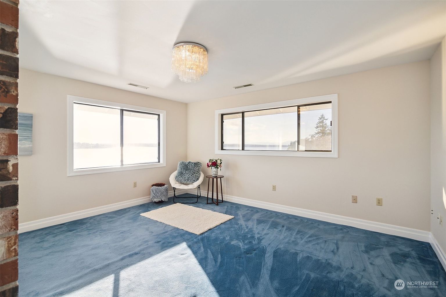 This bright, carpeted room features blue wall-to-wall carpeting and neutral-toned walls, creating a calm atmosphere. Two large windows provide ample natural light and expansive views of the water outside. A decorative chandelier hangs from the center of the ceiling, while a small accent chair and side table are positioned in the corner, offering a cozy seating area.