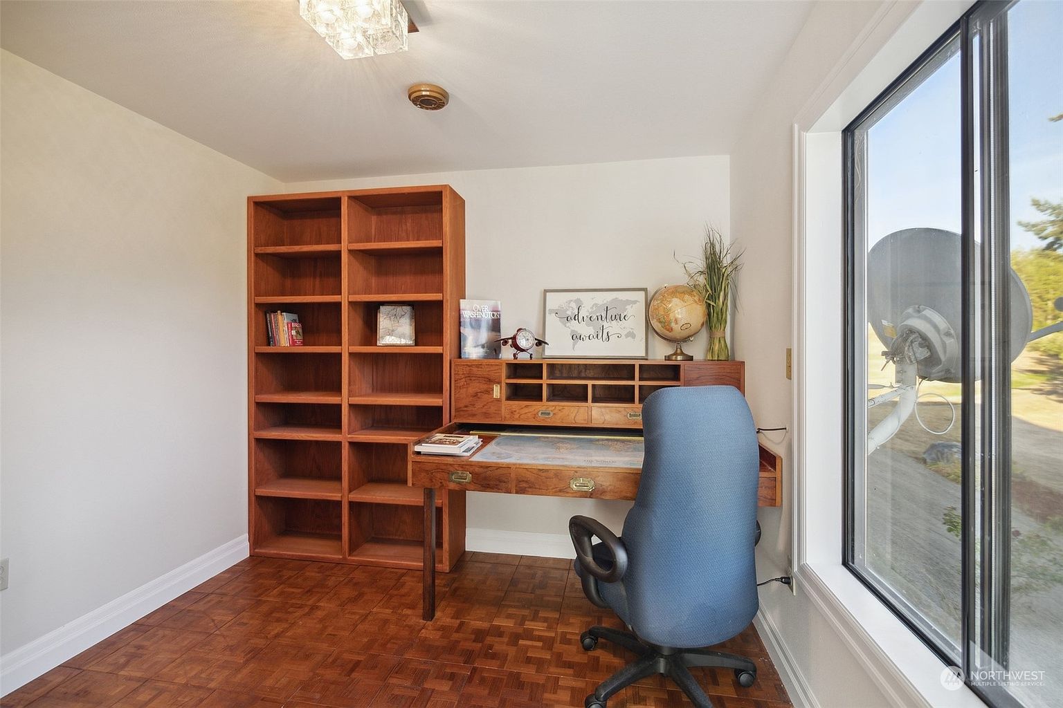 This home office features a classic wooden desk with a hutch and an adjacent tall bookshelf, providing ample storage and workspace. The room is finished with parquet wood flooring and a large window that allows for natural light and a view of the exterior. A blue office chair is positioned at the desk, creating a functional and inviting study area.