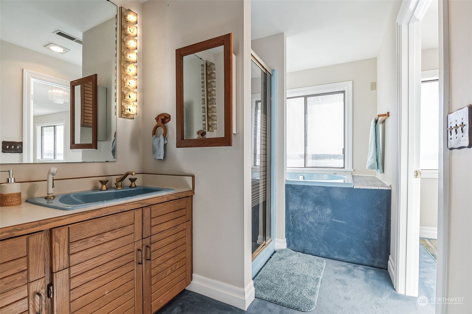 This bathroom features a vanity with a blue sink and light wood cabinetry, paired with a matching blue-tiled soaking tub area. A glass-enclosed shower sits between the vanity and the tub, which is positioned beneath a window that provides natural light. The space is finished with blue carpeting and a wall-mounted light fixture above the mirror, creating a cohesive, vintage-inspired aesthetic.
