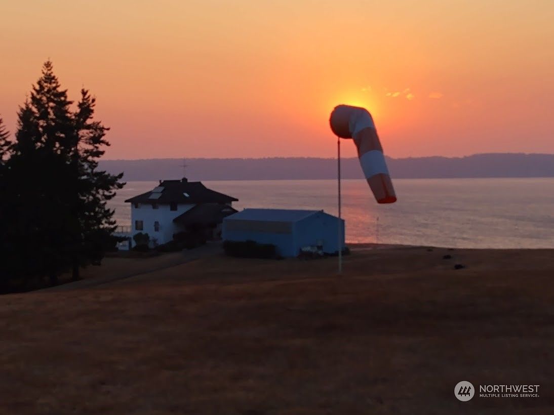 This aerial perspective captures a coastal property at sunset, featuring a white house and a separate blue outbuilding situated on a grassy lot overlooking the water. A prominent windsock stands in the foreground, silhouetted against the vibrant orange sky as the sun dips toward the horizon. The scene offers a serene, expansive view of the shoreline and the tranquil sea beyond.