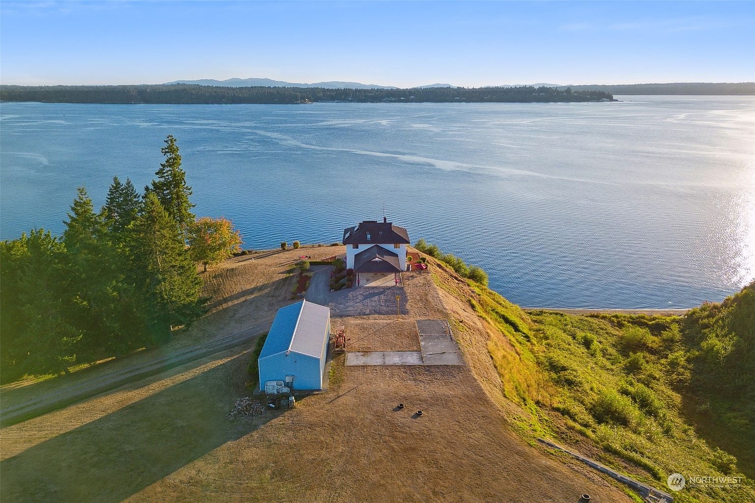 This high-angle aerial view captures a secluded waterfront property situated on a prominent bluff overlooking a vast, calm bay. The site features a main residence with a covered entryway, a detached blue metal outbuilding, and a large, open grassy lot that extends toward the shoreline. The expansive water views and the dramatic cliffside positioning create a serene and private atmosphere, highlighting the unique coastal landscape.