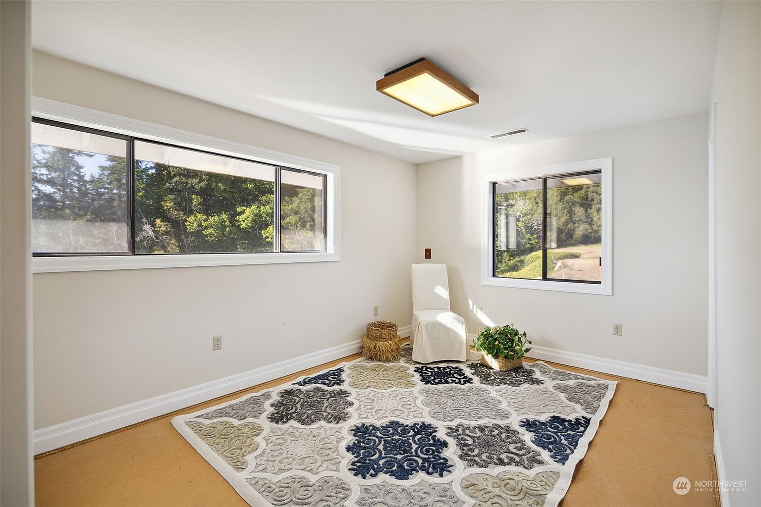 This bright, neutral-toned room features large windows on two walls that provide ample natural light and views of the surrounding trees. The space is anchored by a large, patterned area rug and includes a single chair and a small potted plant, offering a minimalist aesthetic. The room has light-colored walls and a simple, clean layout suitable for a home office or quiet sitting area.