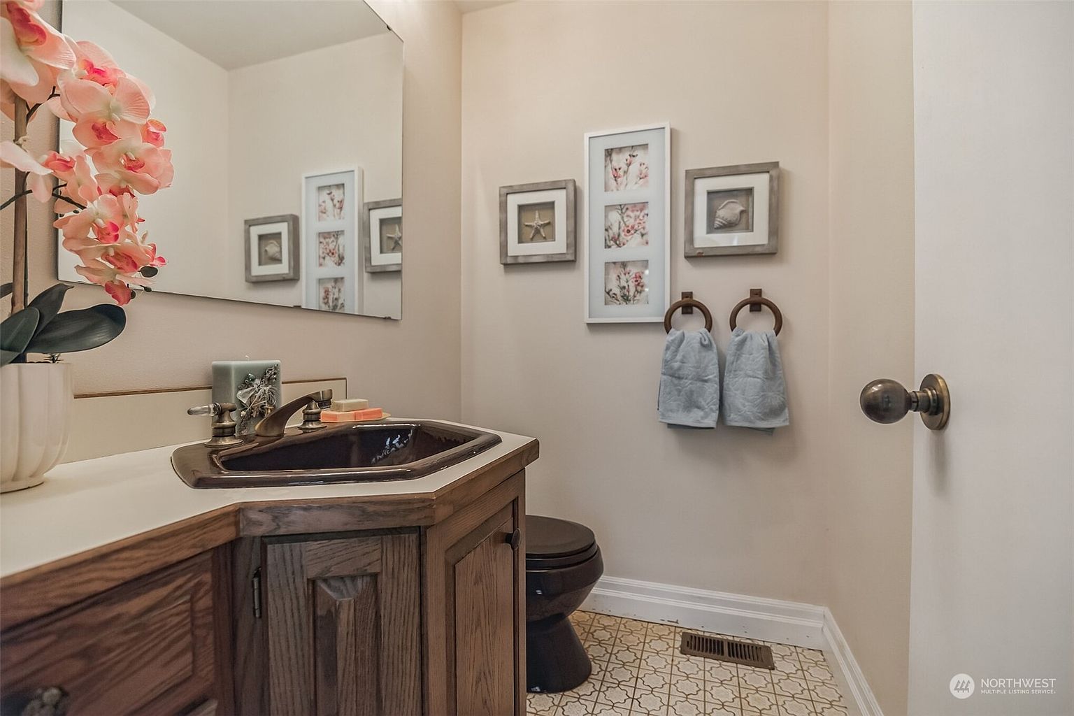 This compact powder room features a dark-toned sink and toilet, contrasting with the light-colored walls and patterned floor tile. A wooden vanity cabinet provides storage, while a mirror above the sink reflects the wall decor and artificial floral arrangement. The space is accented with framed wall art and two light blue hand towels hanging on circular rings.