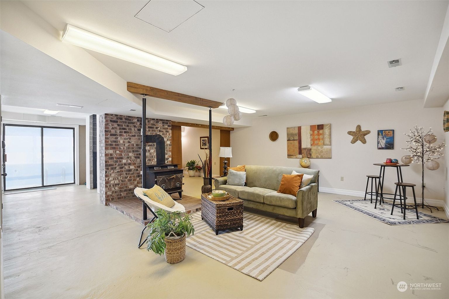 This spacious basement living area features a neutral color palette and a prominent brick chimney breast housing a wood-burning stove. The room is furnished with a comfortable sofa, a woven storage ottoman, and a small dining set, creating a versatile multi-use space. Large sliding glass doors on the left provide natural light, while the open layout and exposed structural beam add to the casual, rustic aesthetic.