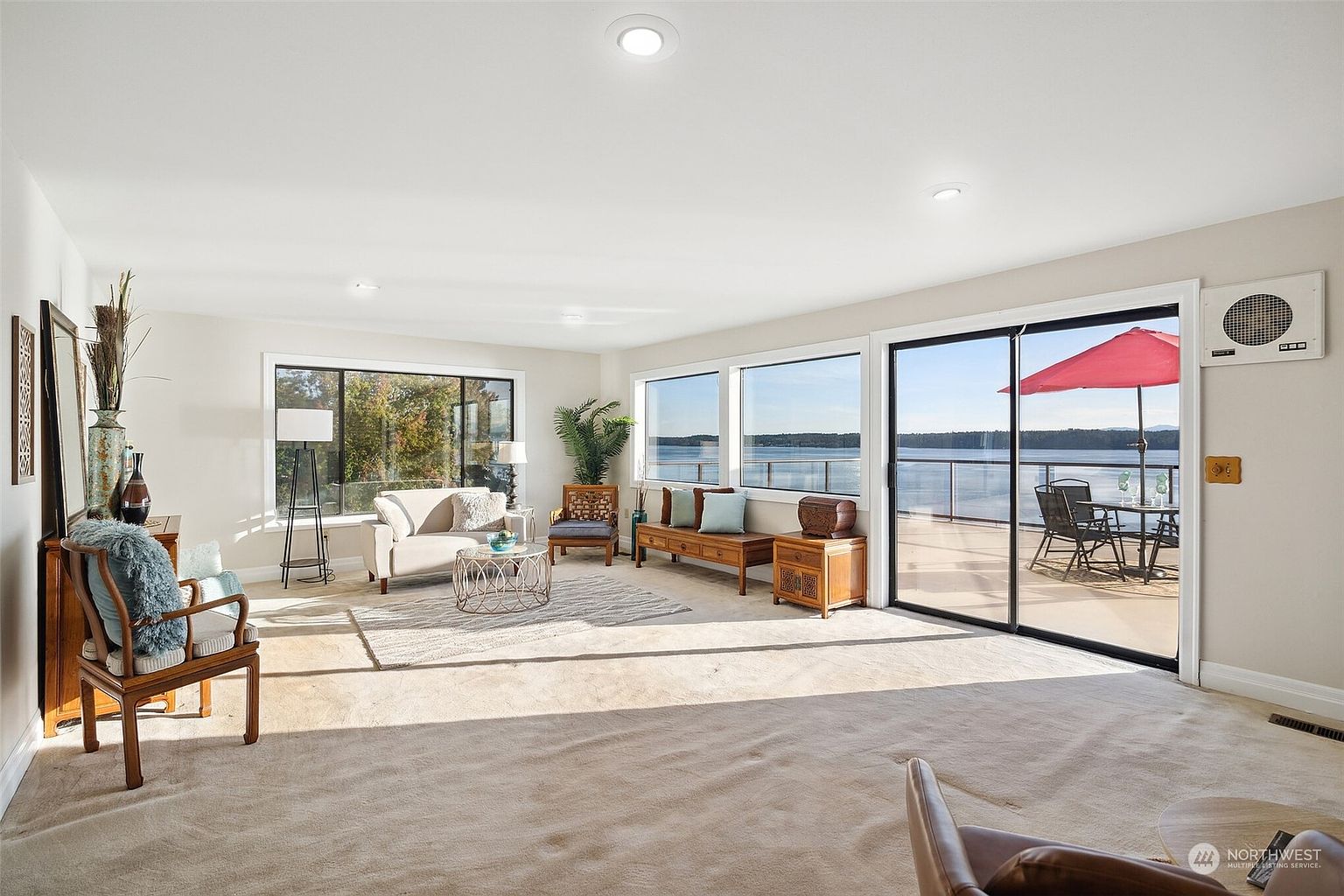 This bright and airy living room features neutral carpeting and a spacious layout, centered around a coffee table and sofa. Large windows and a sliding glass door provide expansive views of the water and access to an outdoor deck, filling the space with natural light. The room is decorated with a mix of wood furniture and comfortable seating, creating a welcoming and serene atmosphere.